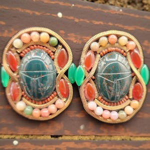 May include: Ornate, vintage-style jewelry piece featuring two symmetrical, gold-toned frames. Each frame has a central, dark green scarab beetle design, surrounded by colorful beads and gemstones in shades of red, orange, and green. The piece is set against a dark brown background.