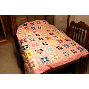Puede incluir: Un edredón hecho a mano con un fondo rosa y un diseño de patchwork. El edredón presenta cuadrados de varios colores y patrones, incluyendo telas florales, a cuadros y lisas. El edredón está sobre una mesa de madera.