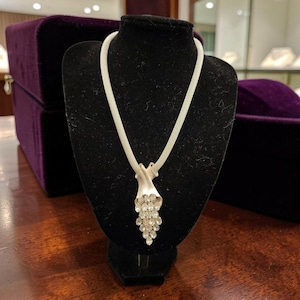 Puede incluir: Un collar plateado con un colgante en forma de flor o campana estilizada, adornado con cristales transparentes y brillantes. El collar se exhibe sobre un busto de terciopelo negro, resaltando su elegante diseño. El fondo incluye una superficie de madera oscura y una caja de terciopelo morado.