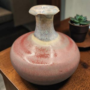 May include: A handcrafted ceramic vase with a bulbous body and a flared, mushroom-shaped top. The vase features a blend of colors, including shades of red, pink, blue, and yellow. The vase is approximately 6 inches tall.