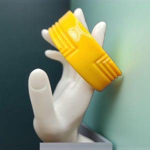 May include: A bright yellow, Art Deco style bangle bracelet. The bracelet features geometric designs and is displayed on a white hand-shaped jewelry holder. The background is a mix of teal and light green.