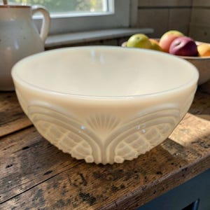 Puede incluir: Cuenco crema de estilo vintage con un patrón decorativo en relieve en el exterior. El cuenco tiene una forma ancha y redondeada y se apoya sobre una superficie de madera. El diseño presenta un motivo en forma de abanico. Una jarra y un frutero están en el fondo.