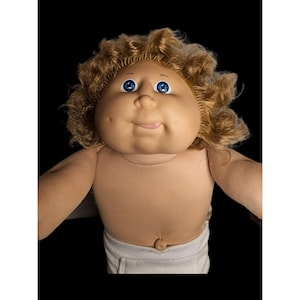 May include: A vintage Cabbage Patch Kid doll with a round face, blue eyes, and blonde curly hair. The doll has a tan fabric body and is wearing white shorts. The doll's tongue is sticking out.
