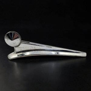 May include: A polished chrome metal hood ornament featuring a sleek, elongated, tapered body with a circular, cone-shaped accent at the front. The object has a smooth, reflective surface and a streamlined design that tapers to a point at the rear.