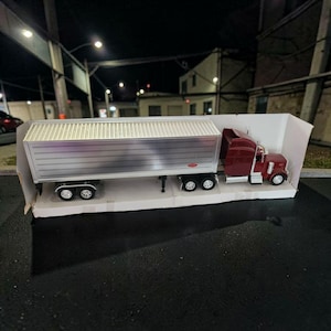 May include: A model semi-truck with a maroon cab and a silver trailer. The trailer has a corrugated design. The truck is displayed in a white cardboard box, with the wheels visible. The scene is set at night.