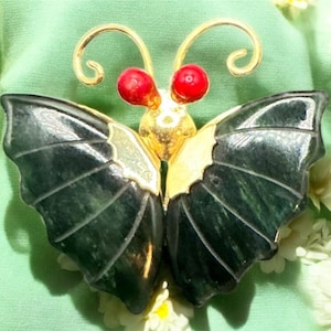 Puede incluir: Broche de mariposa con cuerpo dorado, detalles rojos y alas verde oscuro. Las alas tienen un patrón detallado y veteado. El broche se encuentra sobre un fondo verde claro con flores blancas.