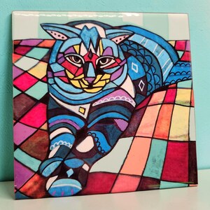 May include: A square tile featuring a geometric cat design. The cat is rendered in blue, purple, red, yellow, and black, set against a backdrop of multi-coloured squares. The artwork has a stained-glass appearance.