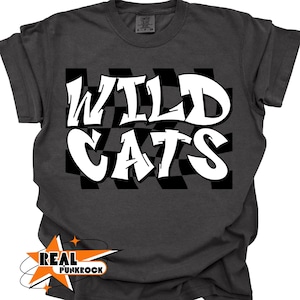May include: Dark gray t-shirt with the words "WILD CATS" in white, stylized graffiti font. The text is set against a black and white checkered background. The shirt has a "REAL PUNKROCK" logo in the lower left corner.