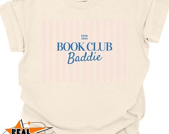May include: Cream-colored t-shirt with a pink and white striped rectangle design. The text "ESTD 2025 BOOK CLUB Baddie" is printed in blue. The shirt is made of a soft, comfortable material.