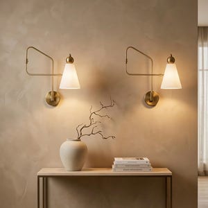 May include: Two wall sconces with brass arms and white cone-shaped shades are mounted on a beige wall. A beige vase with a branch and a stack of books sit on a light wood table. The room has a warm, inviting atmosphere.
