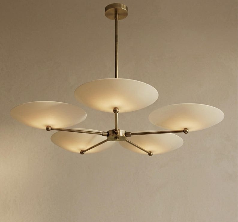 May include: A modern chandelier with five frosted glass shades. The light fixture has a brass finish and a central hub with radiating arms. Each shade is illuminated, casting a warm glow.
