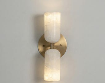 May include: A modern wall sconce with two white, cylindrical light fixtures. The fixtures are mounted on a round, brass-colored backplate. The sconce is designed to provide ambient lighting and has a contemporary aesthetic.