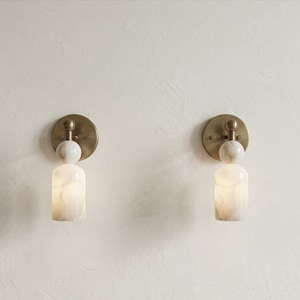 May include: Two wall sconces featuring a circular brass backplate, a spherical white marble accent, and a cylindrical white marble shade. The fixtures emit a soft, warm glow from within the stone, showcasing natural veining patterns in the marble.