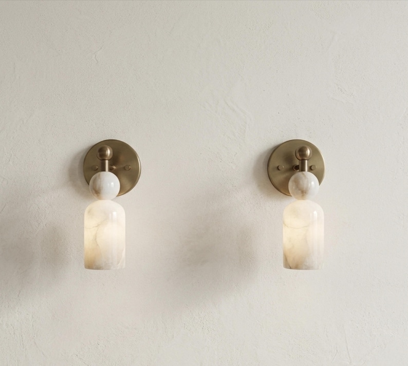 May include: Two wall sconces featuring a circular brass backplate, a spherical white marble accent, and a cylindrical white marble shade. The fixtures emit a soft, warm glow from within the stone, showcasing natural veining patterns in the marble.