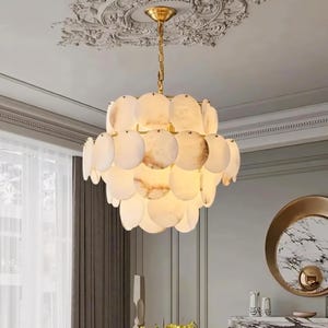 May include: A modern chandelier with a gold chain and canopy. The light fixture features multiple, layered, oval-shaped, off-white and beige alabaster shades. The chandelier hangs in a room with a decorative ceiling and a large round mirror.