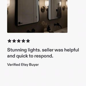 May include: Two wall-mounted light fixtures featuring a spherical white glass shade atop a cylindrical base, positioned on either side of two rectangular mirrors with thin black frames. The lights emit a warm glow against a neutral wall.