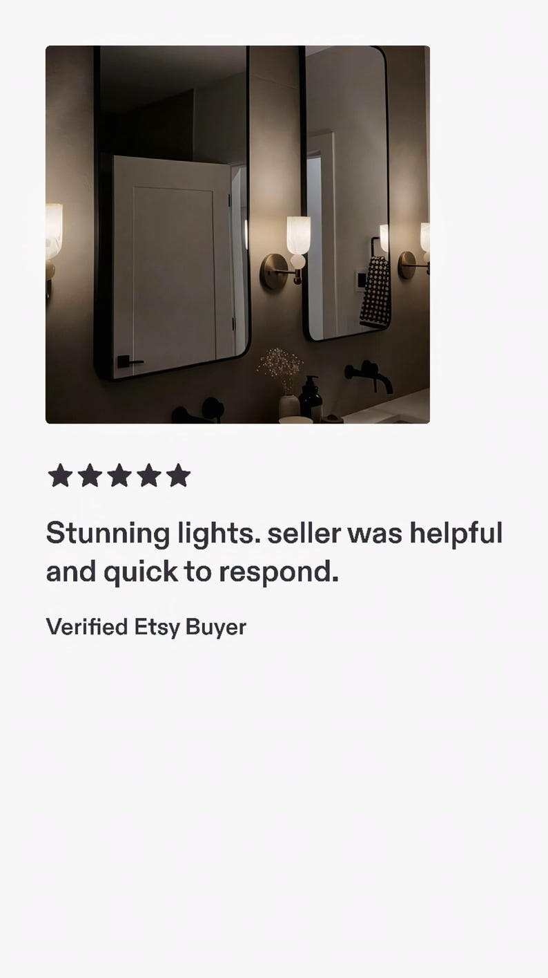 May include: Two wall-mounted light fixtures featuring a spherical white glass shade atop a cylindrical base, positioned on either side of two rectangular mirrors with thin black frames. The lights emit a warm glow against a neutral wall.