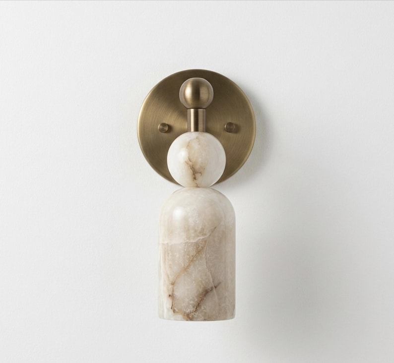 May include: A wall sconce featuring a circular brushed brass backplate with a central vertical arrangement of a small brass sphere, a white marble sphere, and a larger cylindrical white marble base with brown veining throughout the stone.