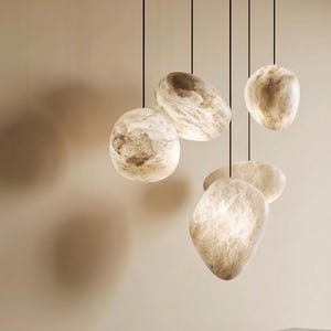Pode incluir: Um conjunto de cinco candeeiros suspensos por fios pretos finos. Cada candeeiro tem uma forma irregular semelhante a uma pedra, com uma superfície translúcida de cor creme e bege marmoreada que brilha, projetando sombras suaves.