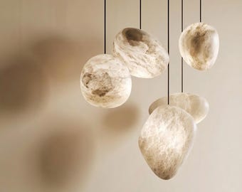 May include: A cluster of five pendant lights suspended by thin black cords. Each light features an irregular, stone-like shape with a translucent, marbled cream and beige surface that glows from within, casting soft shadows on the wall behind.