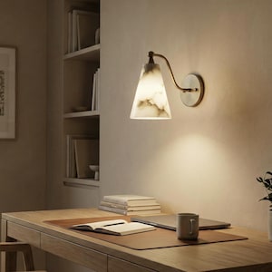 Może przedstawiać: Lampa ścienna z białym i szarym kloszem o marmurowym wyglądzie oraz mosiężnym ramieniem. Oprawa jest zamontowana na beżowej ścianie nad drewnianym biurkiem. Lampa biurkowa zapewnia miękkie oświetlenie obszaru biurka. Biurko ma skórzaną podkładkę, laptopa i kubek.