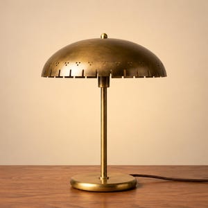 May include: A brass table lamp featuring a dome-shaped shade with a perforated edge pattern and a vertical stem attached to a circular base. The metal surface has a brushed finish, and a dark brown power cord extends from the base to the right.