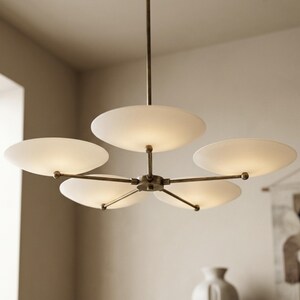 May include: A modern, brass-toned chandelier with five frosted, disc-shaped shades. The light fixture is suspended from a single rod and features a central hub with radiating arms. The shades emit a soft, diffused light.