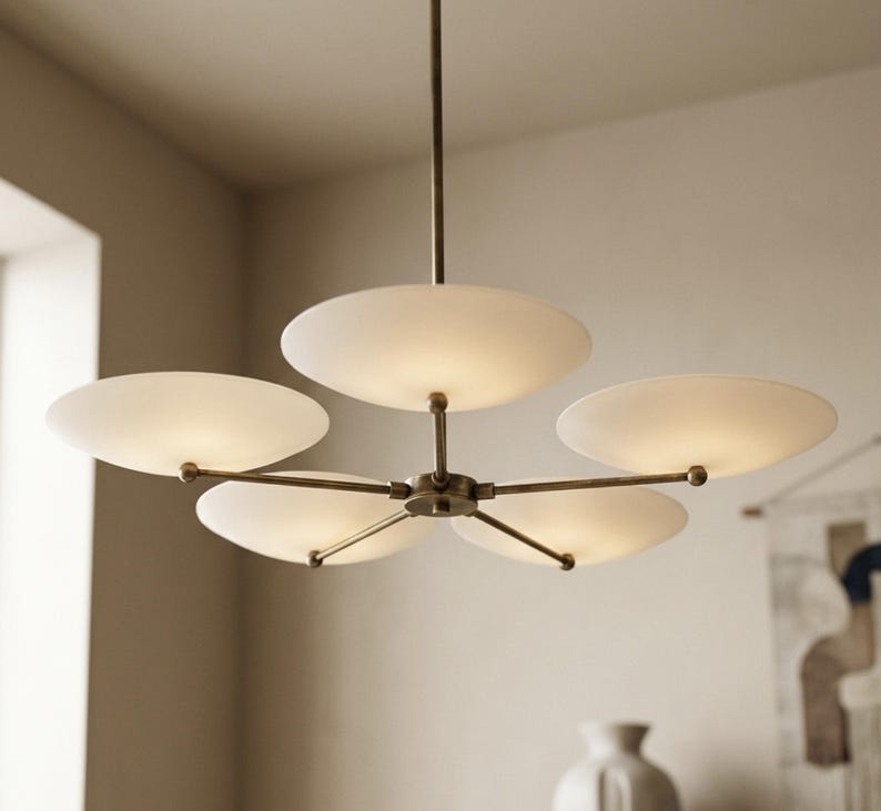 May include: A modern, brass-toned chandelier with five frosted, disc-shaped shades. The light fixture is suspended from a single rod and features a central hub with radiating arms. The shades emit a soft, diffused light.
