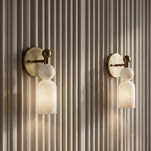 May include: Two wall sconces featuring a circular brushed brass backplate and a horizontal arm connected to a spherical white stone accent. Below the sphere is a cylindrical white stone shade that emits a soft, warm glow from within.