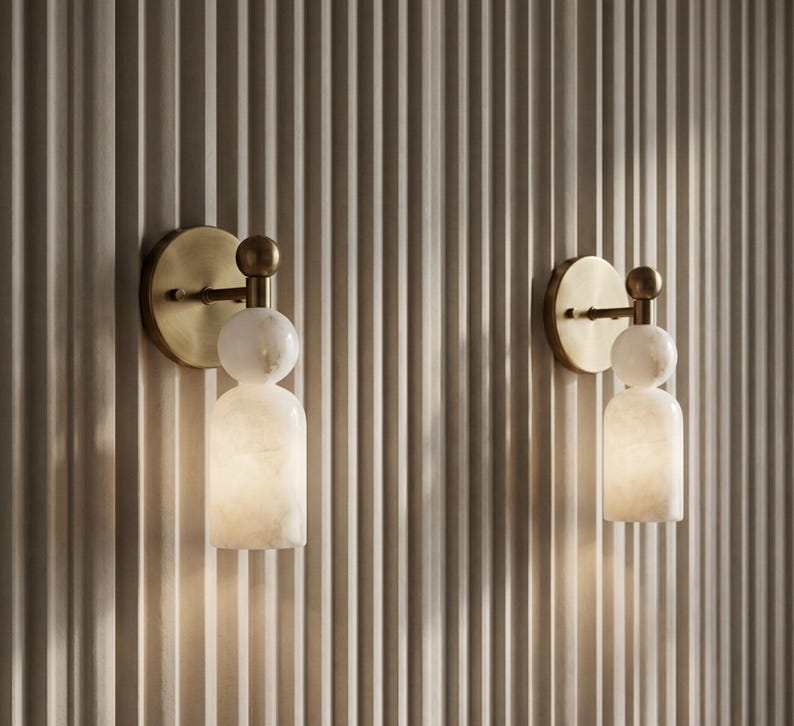 May include: Two wall sconces featuring a circular brushed brass backplate and a horizontal arm connected to a spherical white stone accent. Below the sphere is a cylindrical white stone shade that emits a soft, warm glow from within.