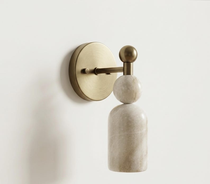 May include: A wall-mounted light fixture featuring a circular brushed brass backplate and a horizontal arm. The fixture includes a spherical brass finial atop a stacked design consisting of a white marble sphere and a cylindrical white marble base.