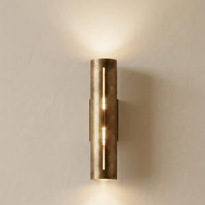 Peut inclure: Applique murale cylindrique avec une finition en laiton chaud. La lampe possède deux fentes verticales qui émettent une lumière douce et chaleureuse. Design minimaliste et moderne, pour l'éclairage intérieur.