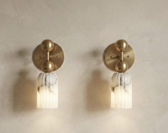 May include: Two wall sconces with a brass finish and a marble-like base. Each sconce has a circular backplate, two spherical accents, and a cylindrical, ribbed shade. The sconces are mounted on a neutral-colored wall.
