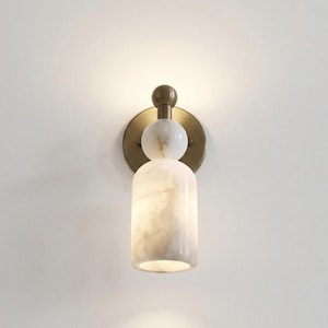 May include: A wall-mounted light fixture featuring a circular brass backplate, a small brass sphere, a white marble sphere, and a cylindrical white marble shade. The lamp emits a soft, warm glow from the top and bottom of the marble cylinder.