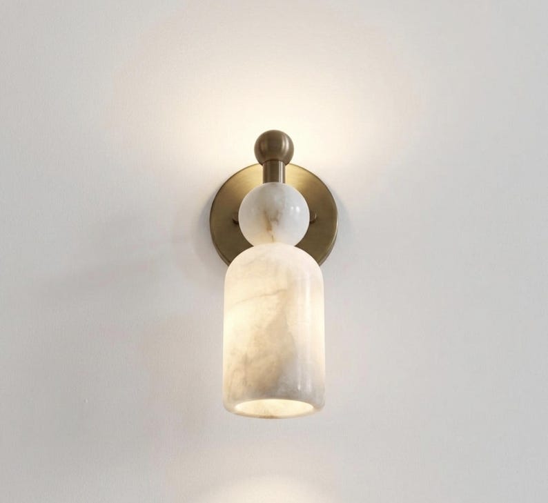 May include: A wall-mounted light fixture featuring a circular brass backplate, a small brass sphere, a white marble sphere, and a cylindrical white marble shade. The lamp emits a soft, warm glow from the top and bottom of the marble cylinder.