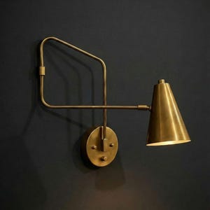 May include: A brass wall sconce with a conical shade and a rectangular arm. The sconce is mounted on a circular base and has a warm, inviting glow. The fixture is made of brass and is mounted on a dark wall.