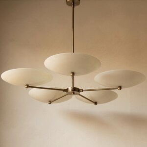 May include: A modern chandelier with five frosted glass disc shades. The fixture has a brass finish and is suspended from a central rod. The shades are arranged in a radial pattern, creating a soft, diffused light.