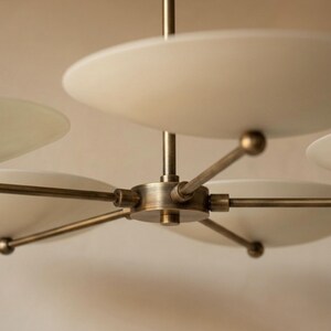 May include: A modern chandelier with a brass finish and multiple white, disc-shaped shades. The fixture has a central hub with radiating arms, each supporting a shade. The design is minimalist and elegant, suitable for various interior styles.
