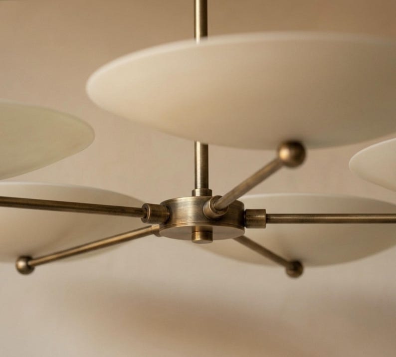 May include: A modern chandelier with a brass finish and multiple white, disc-shaped shades. The fixture has a central hub with radiating arms, each supporting a shade. The design is minimalist and elegant, suitable for various interior styles.