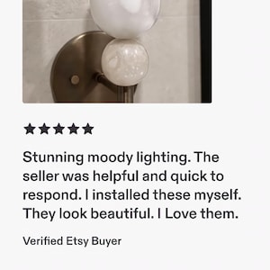 May include: A wall-mounted light fixture featuring a circular bronze-toned metal base and a vertical cylindrical glass shade. A small, spherical, textured white glass orb sits between the base and the main cylindrical glass light component.