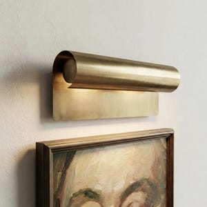 May include: A brass wall sconce with a curved, cylindrical shade and a rectangular backplate. The sconce is mounted on a white wall above a framed painting with a portrait.