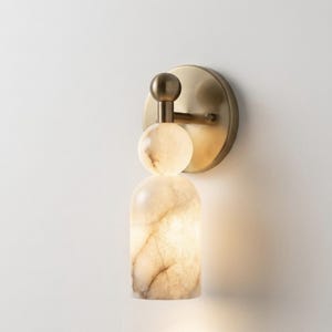 May include: A wall sconce featuring a circular brushed brass backplate and a vertical arm supporting a spherical stone accent above a cylindrical stone shade. The stone components exhibit a translucent, marbled cream and beige veined pattern.