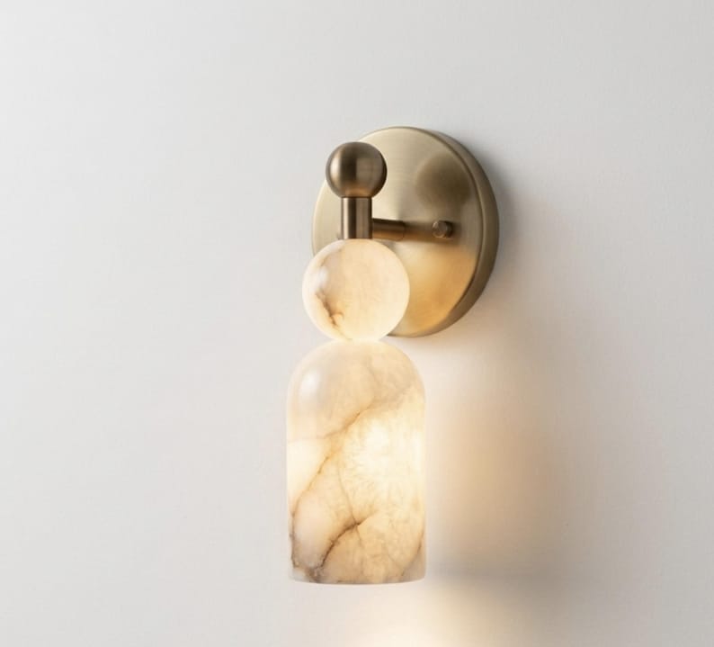 May include: A wall sconce featuring a circular brushed brass backplate and a vertical arm supporting a spherical stone accent above a cylindrical stone shade. The stone components exhibit a translucent, marbled cream and beige veined pattern.