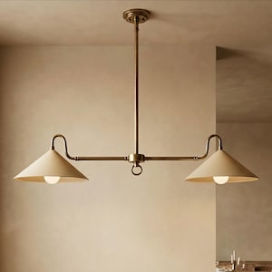May include: A brass pendant light with two cream-coloured, cone-shaped shades. The light fixture has a central vertical rod, horizontal arms, and a ring detail. Each shade houses a single light bulb.