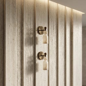 May include: Two wall-mounted light fixtures featuring a circular bronze base, a small spherical accent, and a cylindrical white stone shade. The fixtures are mounted vertically on a textured, light-colored, vertically paneled wall surface.