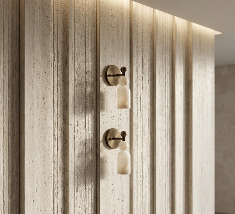May include: Two wall-mounted light fixtures featuring a circular bronze base, a small spherical accent, and a cylindrical white stone shade. The fixtures are mounted vertically on a textured, light-colored, vertically paneled wall surface.