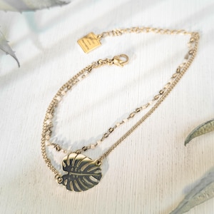 Può includere: Bracciale dorato a più strati con un ciondolo a forma di foglia di Monstera. Il bracciale ha tre delicate catene, una con piccole perline. Una piccola etichetta rettangolare è attaccata alla catena. Il bracciale è esposto su una superficie bianca.