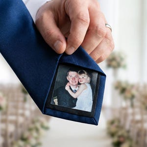 Może przedstawiać: Granatowy krawat z ukrytym zdjęciem mężczyzny i dziecka. Krawat jest podniesiony, odsłaniając obraz w środku. Tło przedstawia ceremonię ślubną z krzesłami i kompozycjami kwiatowymi.