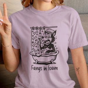 May include: A lilac t-shirt with a black and white cartoon wolf in a bathtub. The wolf, wearing a shower cap, has an angry expression. The text "Fangs in foam" is printed below the image. The shower curtain has bone shapes.