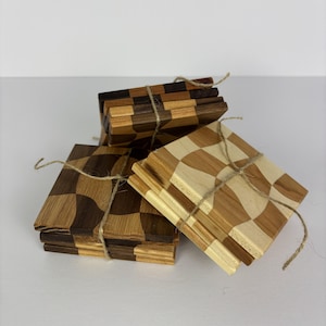 Puede incluir: Juego de posavasos de madera con diseño de tablero de ajedrez. Los posavasos están hechos de diferentes tonos de madera, incluyendo marrones claros y oscuros y beige. Están atados con hilo. Cada posavasos mide aproximadamente 10 cm cuadrados.